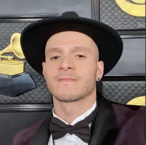 A person wearing a wide-brimmed hat, bow tie, and suit poses in front of Grammy Award-themed decor.
